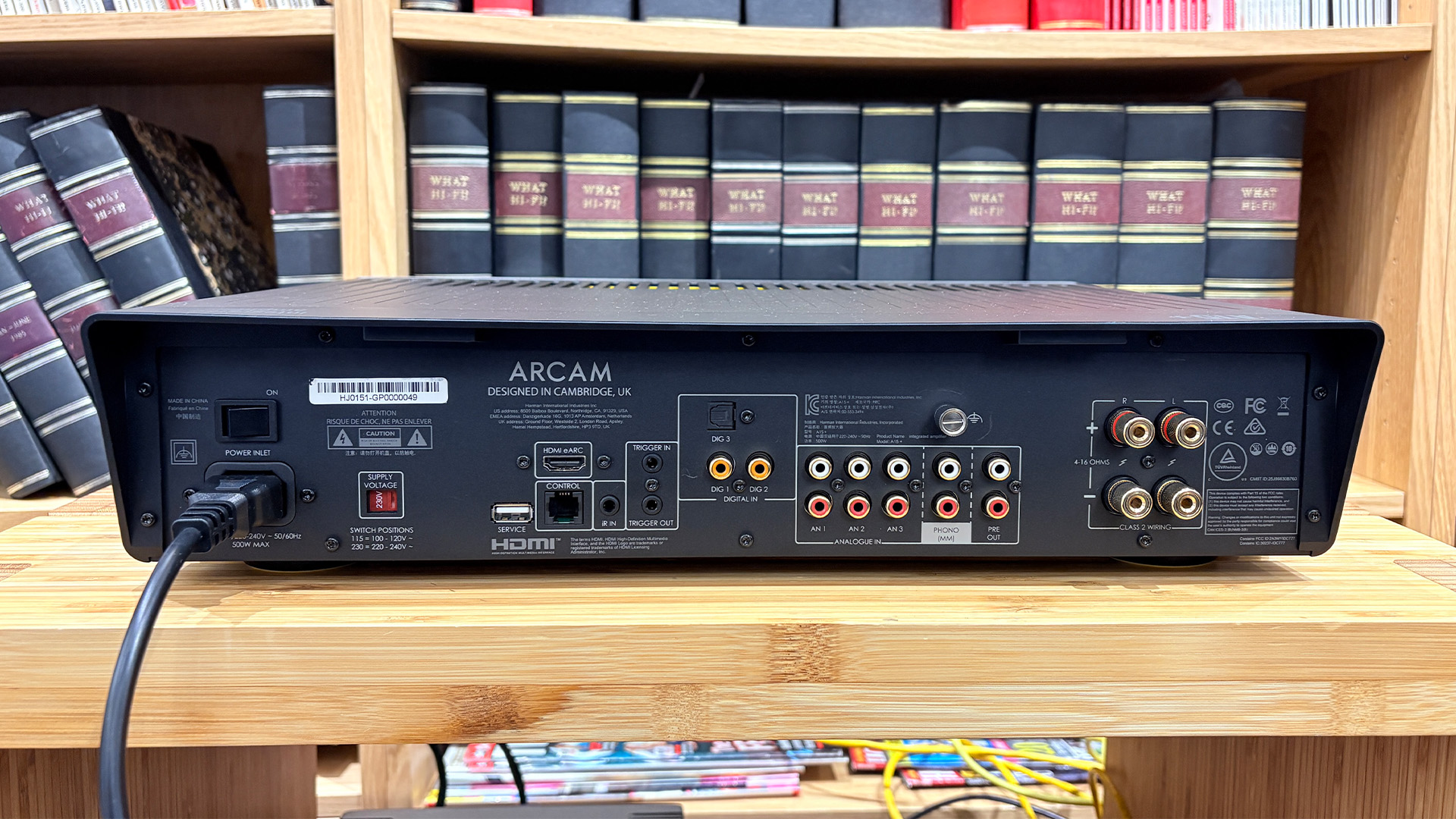 Arcam A15+ integrated amplifier on wooden AV rack in front of bookcase showing rear of unit and connections