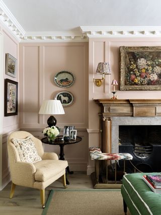 pink living room with vintage paintings, plates on the wall, wall lights, armchair, fireplace, side table and table lamp, bright green footstool, coir and green trimmed rug, wooden floor, fender seat