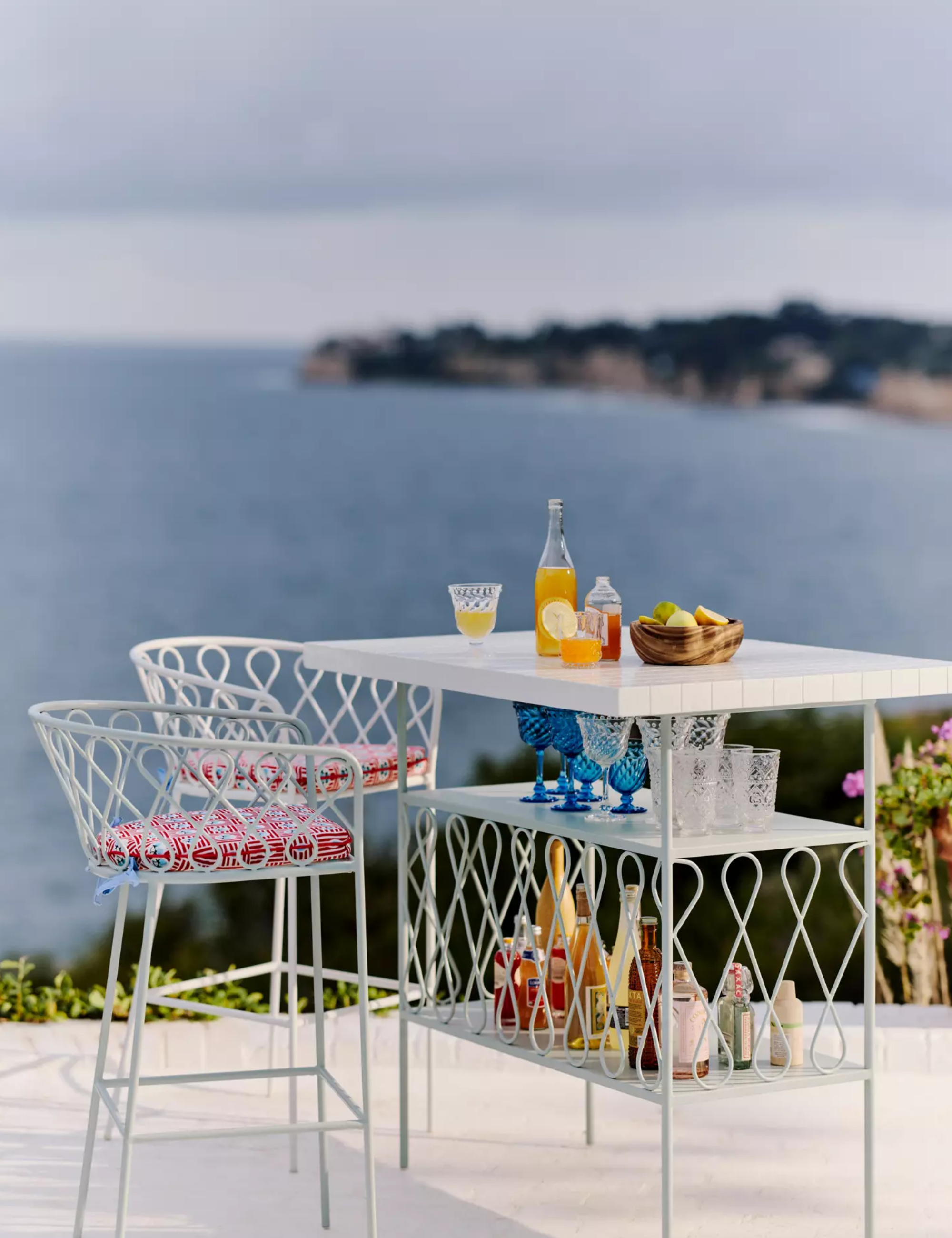 Whimsical, powdery blue South-of-France-style outdoor bar furniture styled beachside with patterned seat cushions and assorted bar accessories.