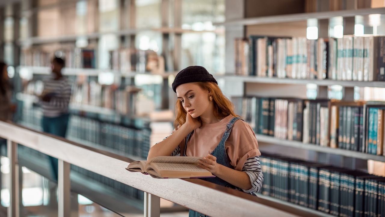 A college student stands at a railing looking over a book in a library.