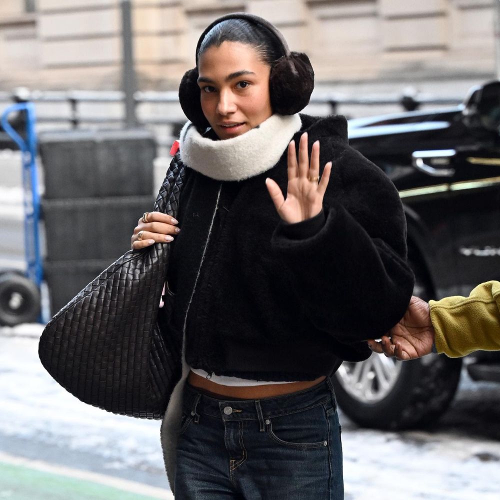Olivia Dean walks outside wearing dark wash low-rise jeans with brown pointed-toe stiletto boots, a black bomber jacket, earmuffs and a Bottega Veneta Maxi Bag.