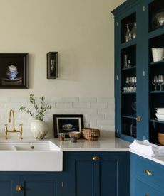 A kitchen with blue cabinetry with brass handles, white walls and work surfaces, with plants and framed artwork and a silver cooker