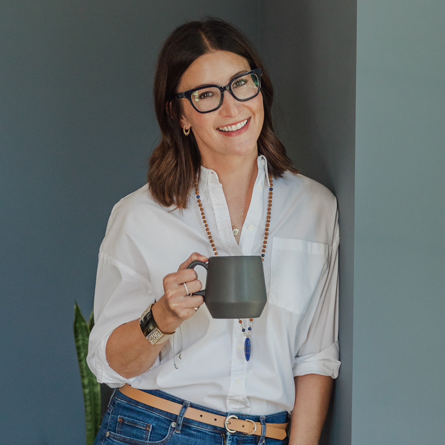 A woman with short brown hair and black framed spectacles in a white shirt with a beaded necklace and blue jeans with a tan belt, holding a green mug