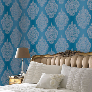 Laurence Llewelyn-Bowen Cote Coutre Damask Wallpaper with an azure blue background and silver damask patter, in a bedroom showing a nicely made bed with elegant headboard and side table with lamp