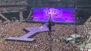 An image from The Eras Tour, London night 1. The stadium is fill and Taylor Swift is disaplyed on a large screen behind a walkway. The view is slightly obstructed by a speaker