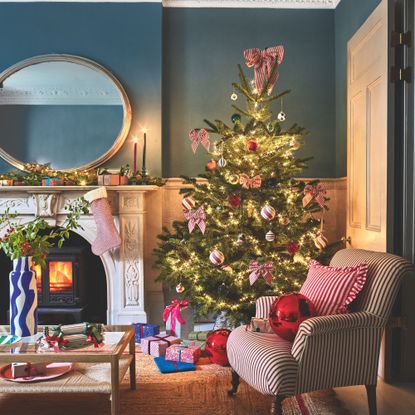 Real christmas tree in a living room next to a log burner and a striped accent chair