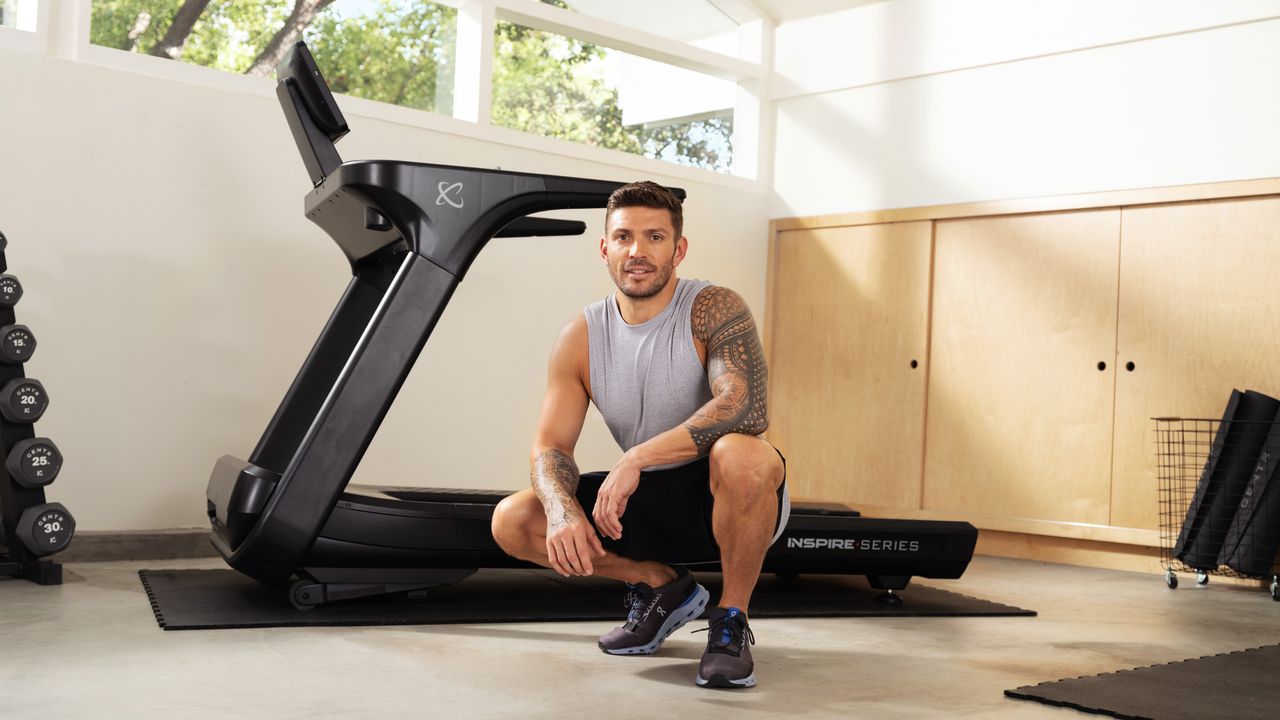 Man crouches in front of treadmill indoors. He is wearing a gray vest, black shorts and black trainers