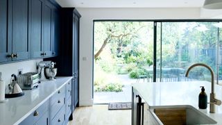 Contemporary blue kitchen with white acrylic worktops looking out through large sliding doors onto garden