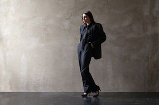 A woman wearing a masculine, oversized black suit in front of an industrial backdrop.