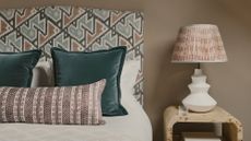 A bed with a patterned fabric headboard, white bedding with two blue throw cushions and a patterned bolster cushion. A wooden side table with a side lamp beside it. 