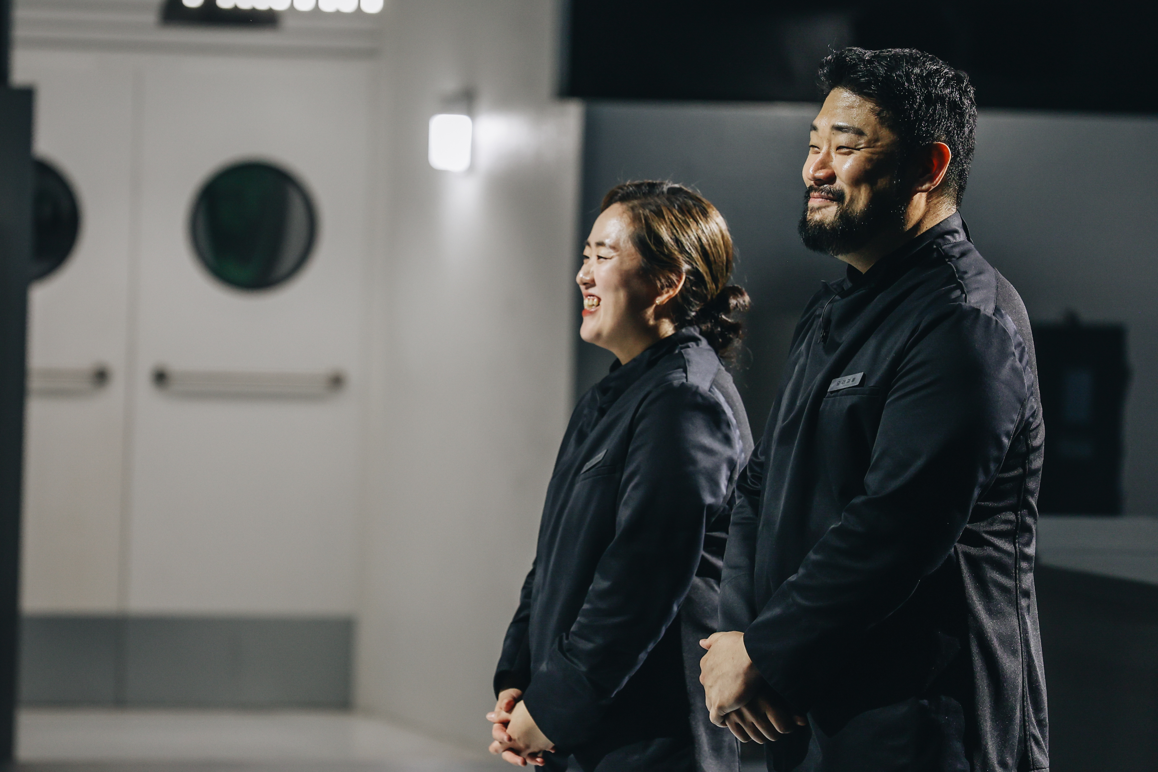 A female chef nicknamed Brewmaster Yun and a male chef nicknamed Culinary Monster stand in an industrial kitchen. They both smile while standing with their hands clasped and wearing all-black uniforms, in 'Culinary Class Wars' season 2.