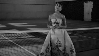 Audrey Hepburn in a white gown with black flowers holding a champaign glass while standing in front of a tennis net.