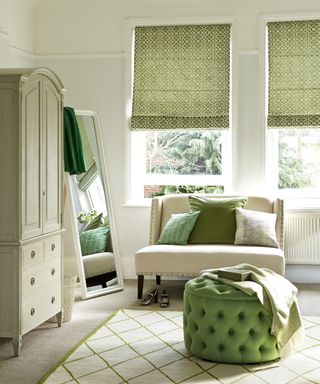 An example of green bedroom ideas showing a white-painted bedroom with green furnishings including printed blinds and an upholstered footstool