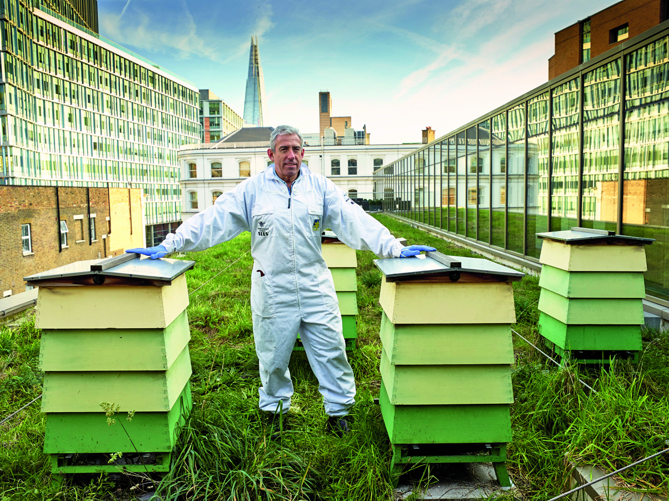 Dale Gibson stands on a roof in London in beekeeping gear with some hives