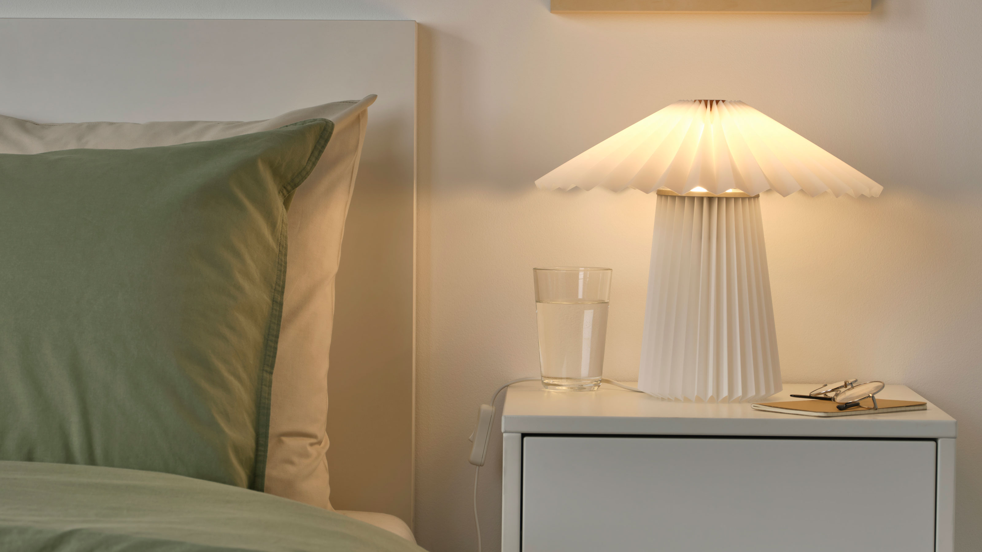 White bedside table next to a bed with pleated table lamp