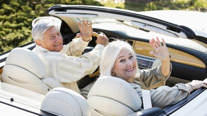 An older couple in a convertible wave goodbye as they drive away.