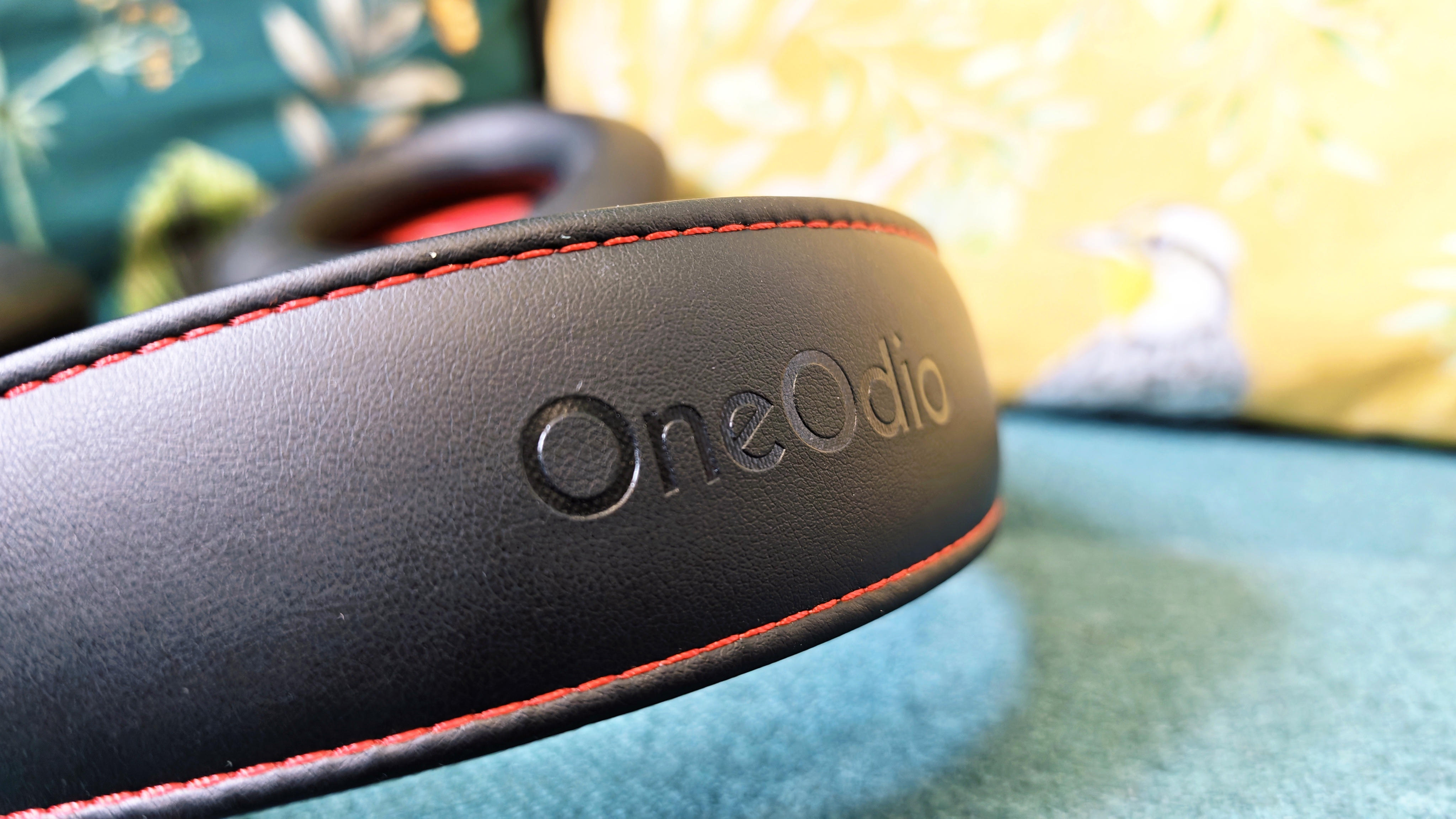 A pair of black OneOdio Studio Max 2 headphones with red internal earcups sitting on a green sofa in front of colourful pillows with birds and flowers on them