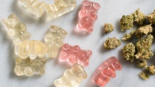 close up of white, orange and pink gummies bears scattered on a table next to clumps of dried cannabis