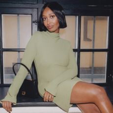 Mona Ali sits on a windowsill wearing a green mini dress with a black top handle bag