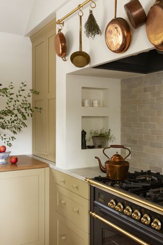 Close-up image of the stove in a beige kitchen design. The stove is black with detailed gold hardware and there is a copper kettle sitting on the burner. There are also copper pots and pans hanging above the stove