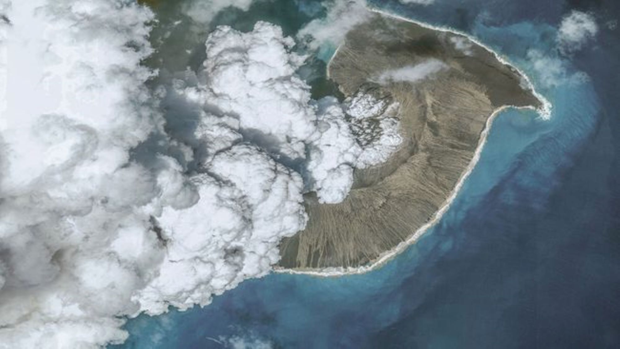 An aerial view of the Tonga volcano eruption in 2022. We see vapor and ash rising from a volcano surrounded by water.