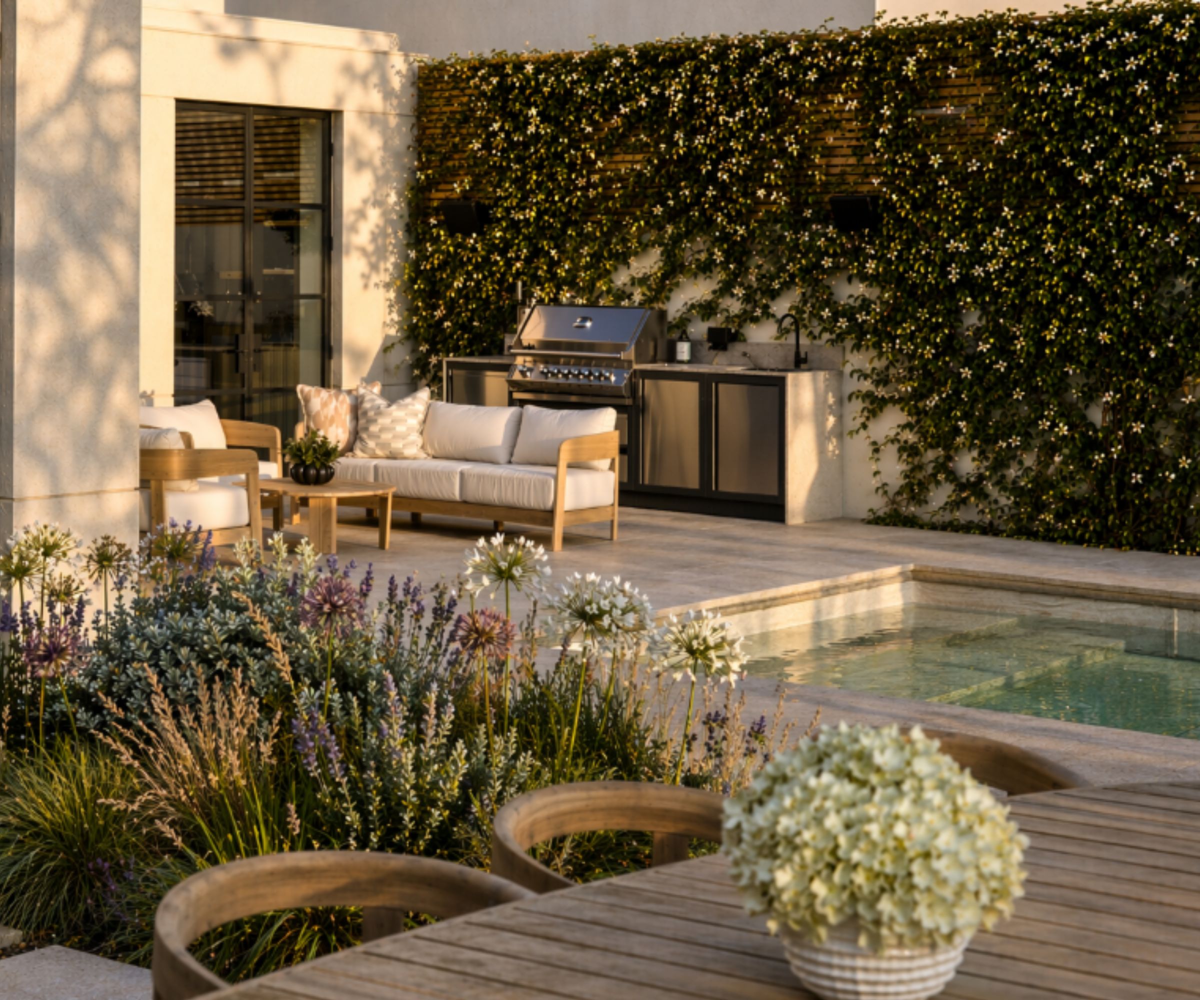 A modular kitchen with a waterfall countertop, star jasmine covering the wall behind it, and a living space in front, all beside a pool