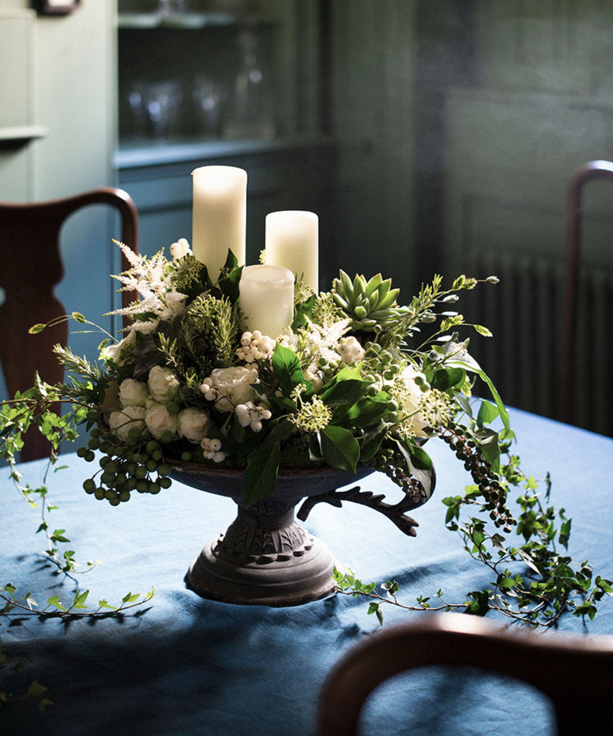 A table centrepiece with greenery and white pillar candles on a blue tablecloth, with dark wood chairs.