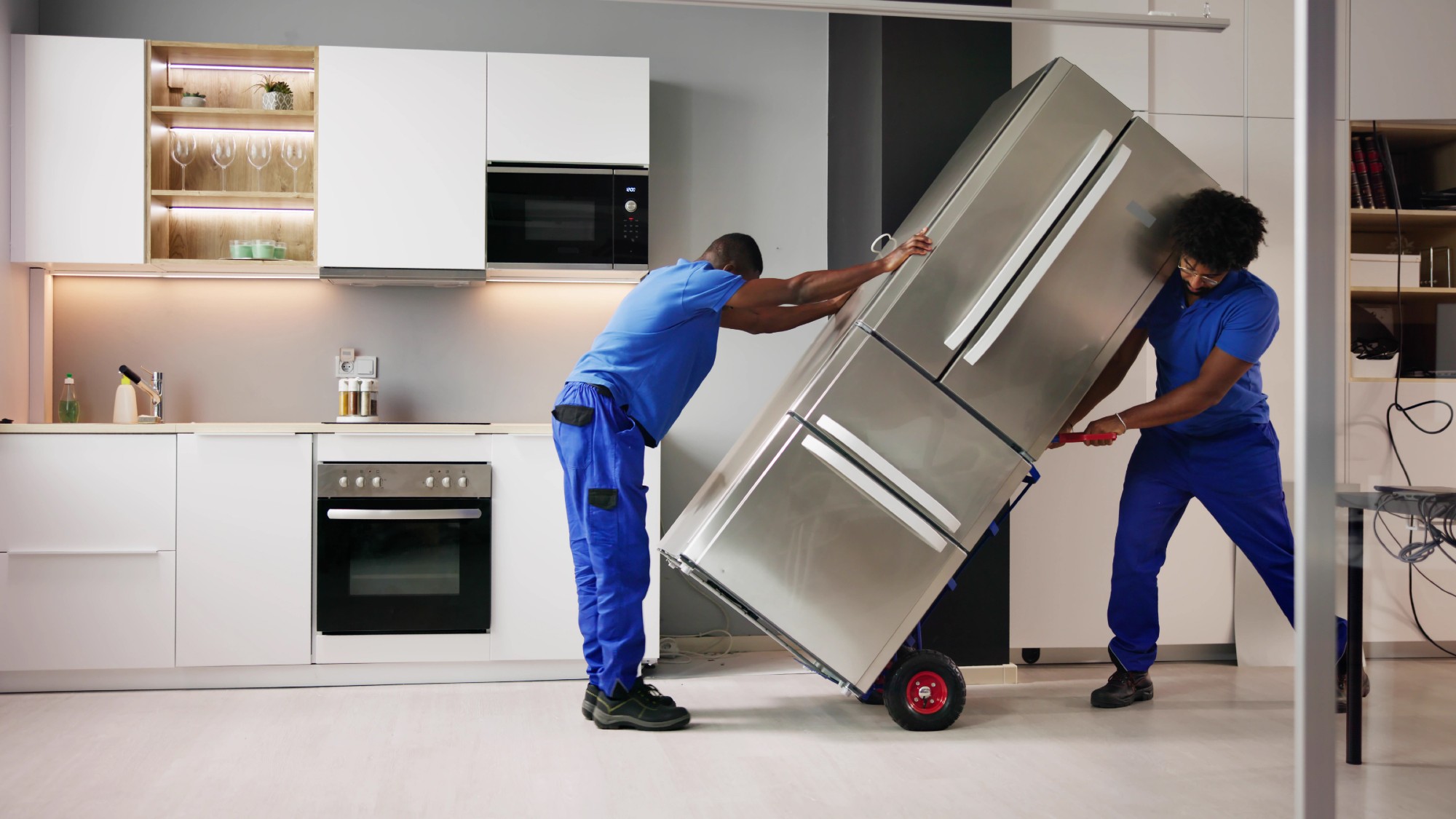Two people wheeling a broken fridge out of a kitchen 