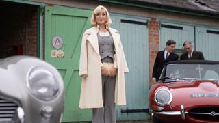 Helen George in a white coat and grey suit stands between two cars in Call the Midwife