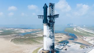 a black and white rocket stands next to a huge metallic launch tower, with the ocean in the background