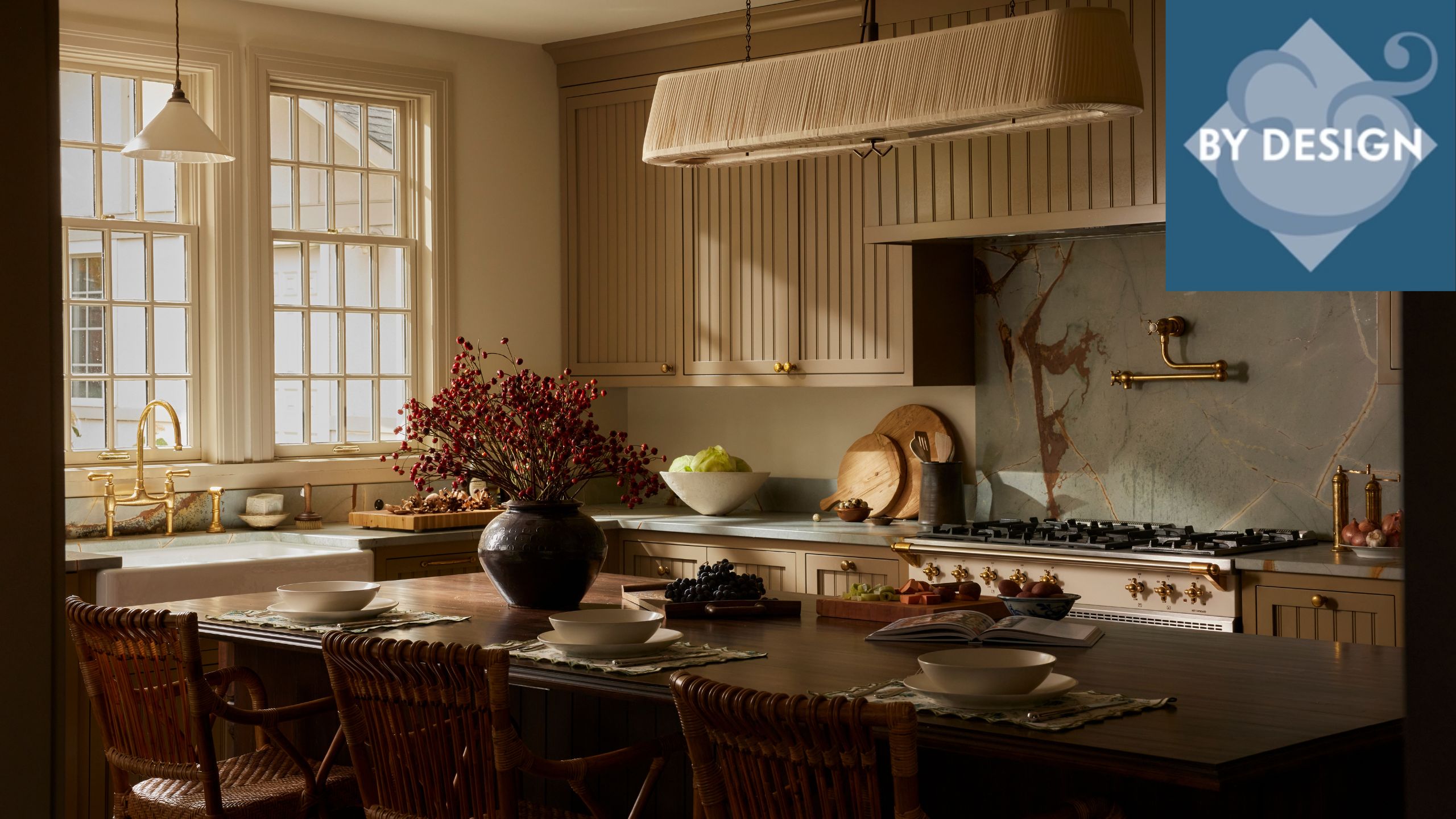 Cozy kitchen, large dark wood dining table in center, decorated with vase of flowers and ableware, slatted wooode kitchen cabinets, stove and windows, sink in background