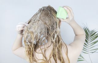 A blonde woman is washing her hair with a green solid shampoo bar