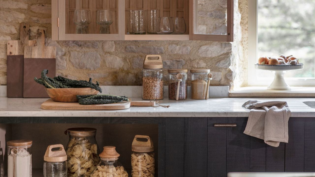 A rustic kitchen with white countertops. Glass jars of dried food goods on the counter and shelves beneath. Dark blue pained cabinets, 