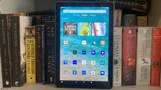 En Amazon Fire HD 10 står lutad mot ett par böcker i en bokhylla med hemskärmen aktiv.