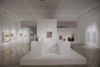 Interior exhibition view at the museum of contemporary art featuring white walls with wall art and white ceiling with silver ceiling lighthing. In the center is a white podium with mini sculptures on display