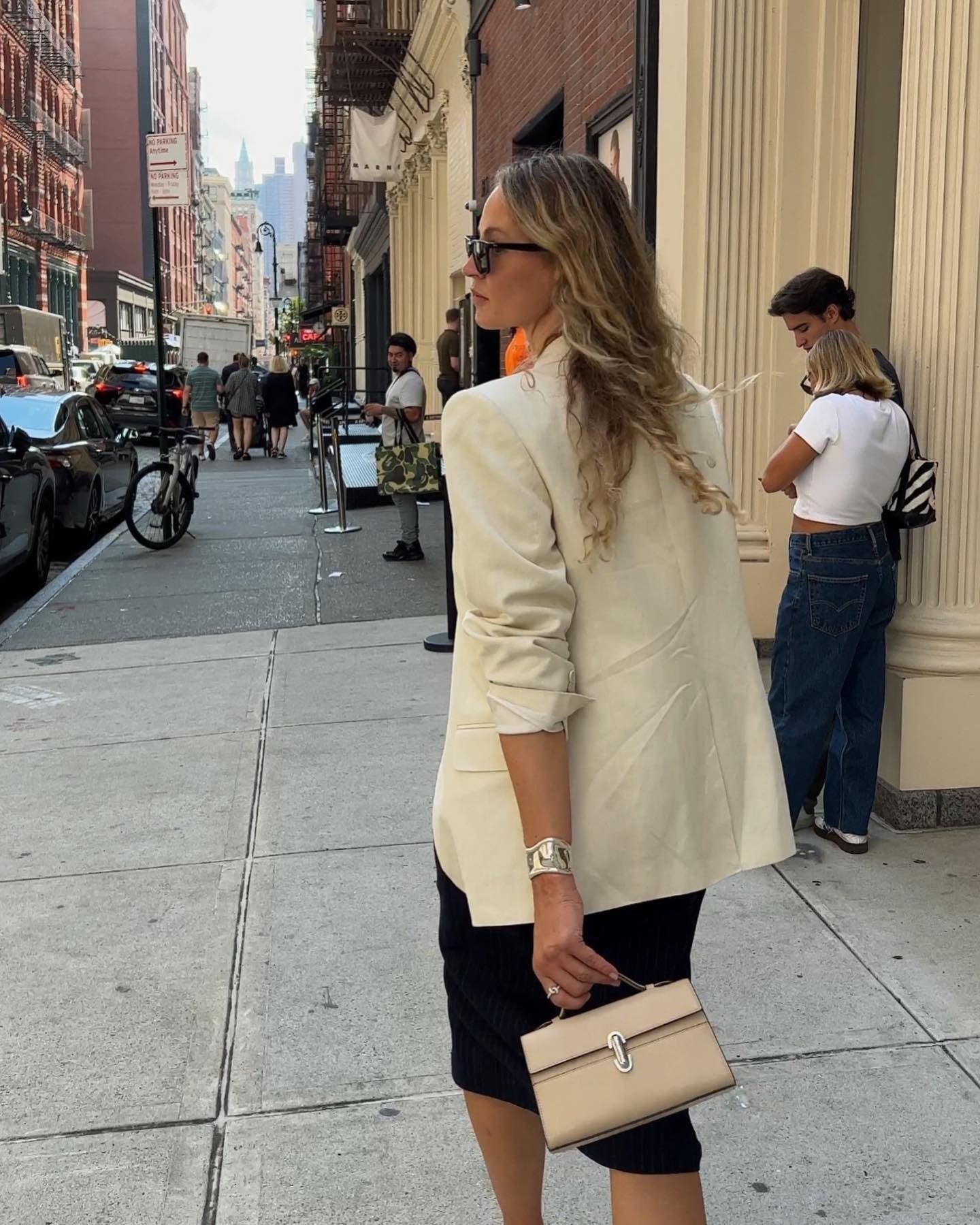 Fashion influencer Anouk Yve walking in Soho NYC wearing black sunglasses, a beige line blazer, black skirt, and tan Savette bag.