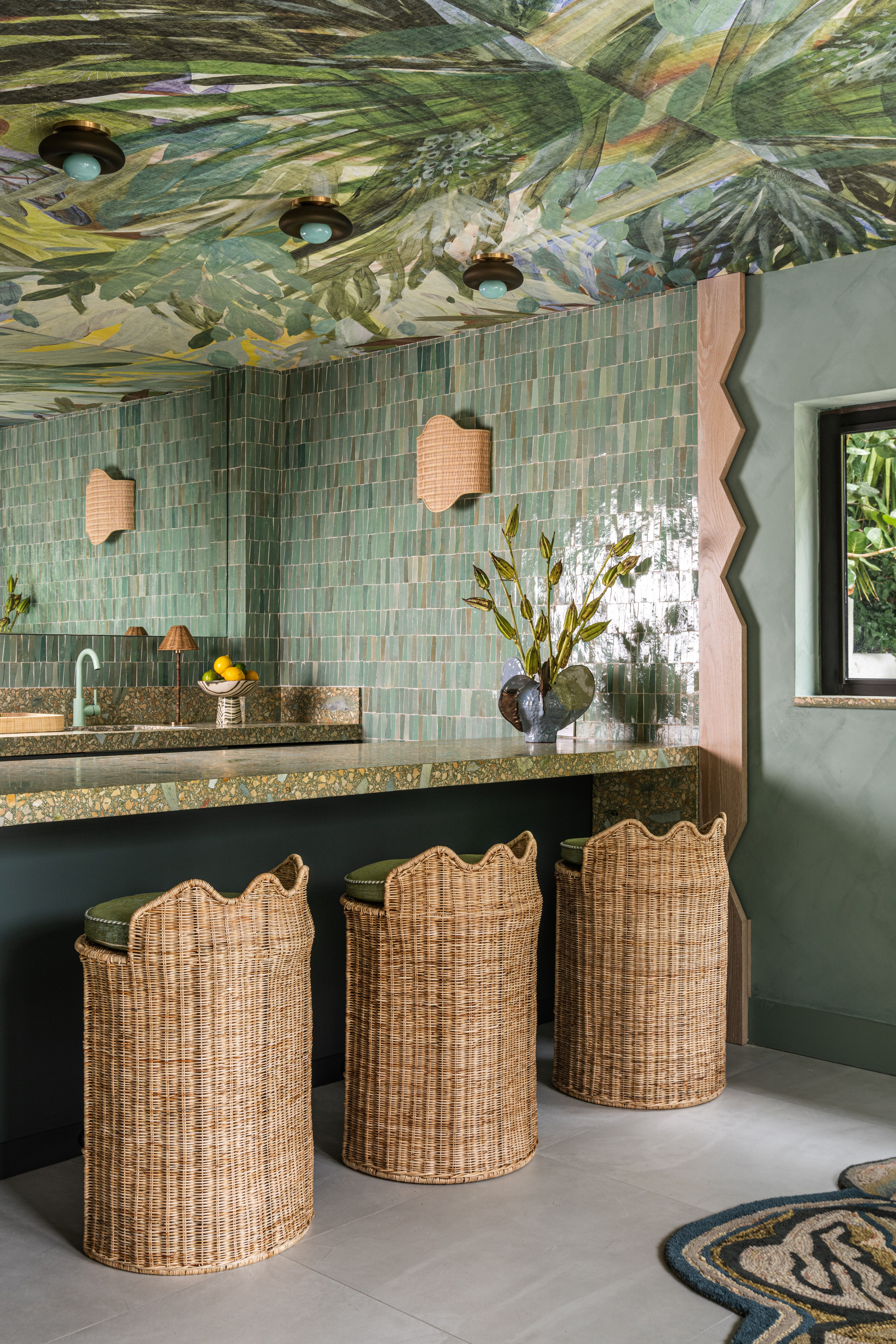 Interior of a tropical home with teal walls, zellige tiles surfaces, wavy, sand-coated details, rattan furniture mixed with bright aquamarine and ochre touches, and a botanical-themed mural ceiling.