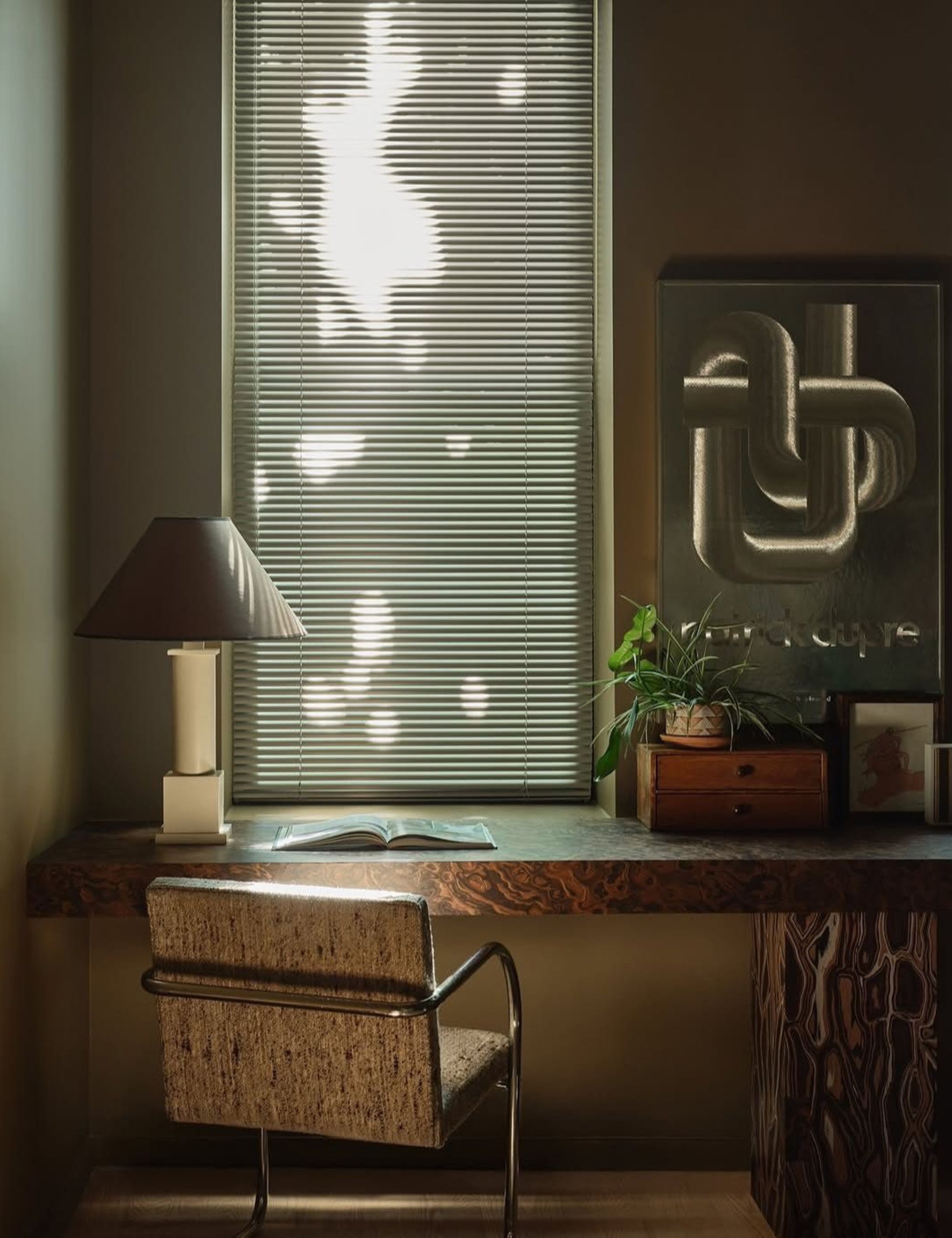 Beige room with dappled light coming in through Venetian blinds, marble desk, with lamp, plant and magazine on top with a silver and grey office chair underneath