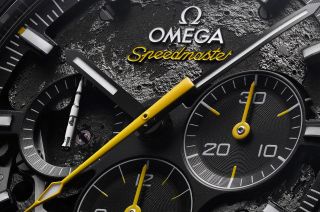 closeup of an omega speedmaster watch face, which has a background photo of the moon's surface and a small white saturn v rocket on one of its dials