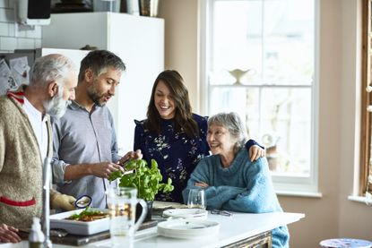 Cheerful mid adult woman with arm around her mother smiling and watching her husband prepare meal, while her mature father with grey hair and beard standing by kitchen island