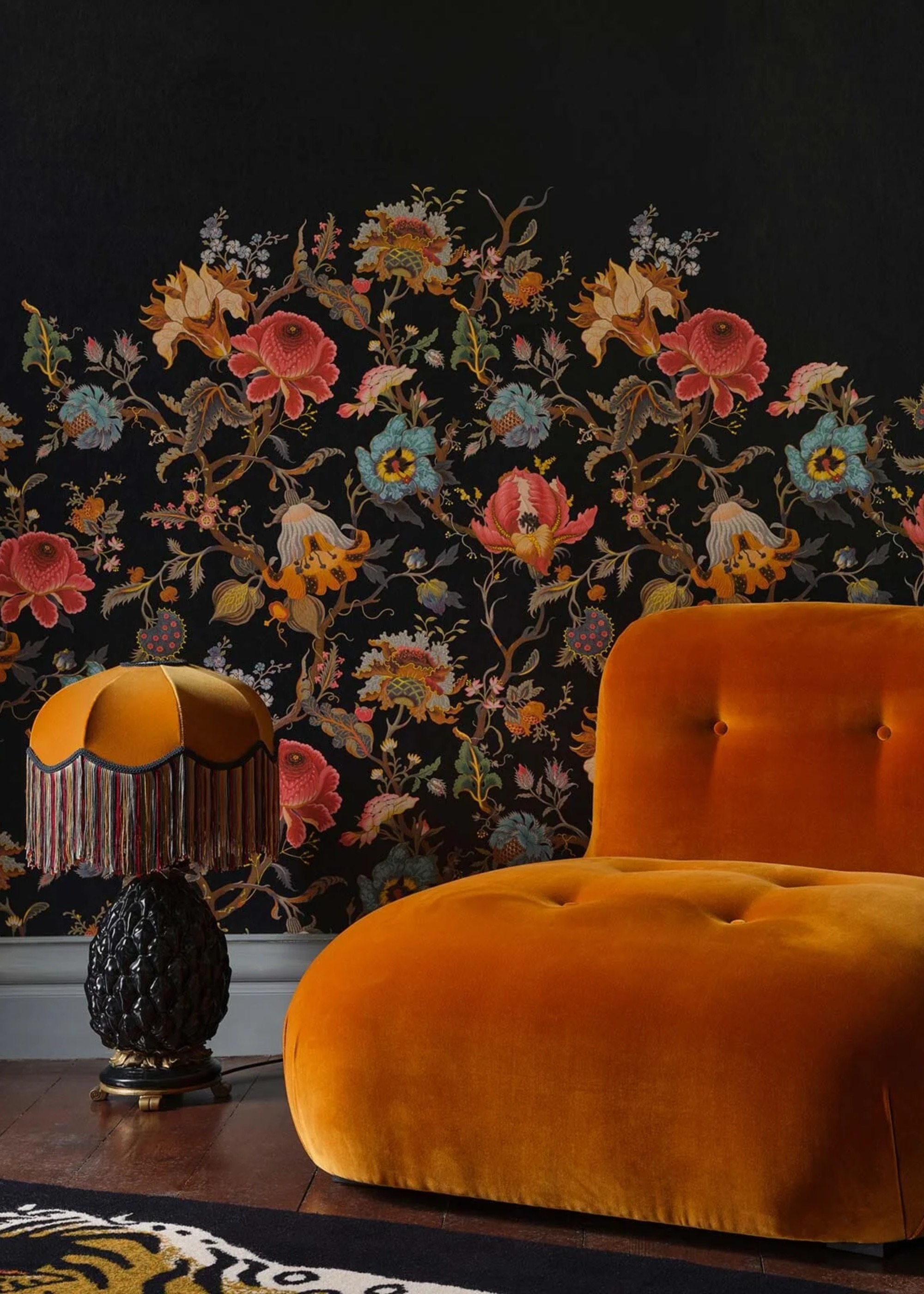A living room with black wallpaper with floral motifs, a table lamp on the floor with a fringe shade, and a deep orange sofa chair