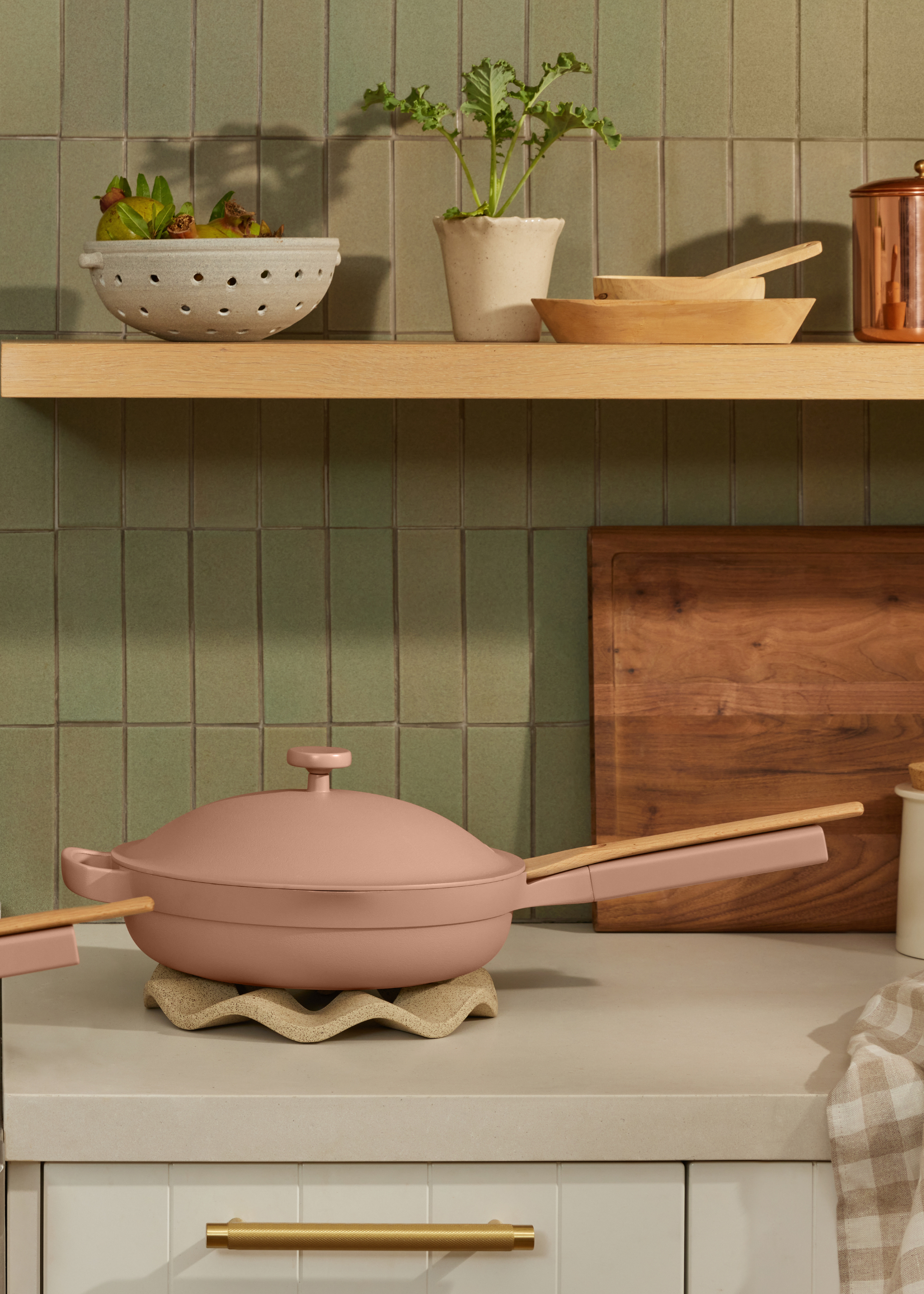 A beige-pink ceramic pan on a ruffled heat rest in a kitchen with floating wood shelves and green tiling