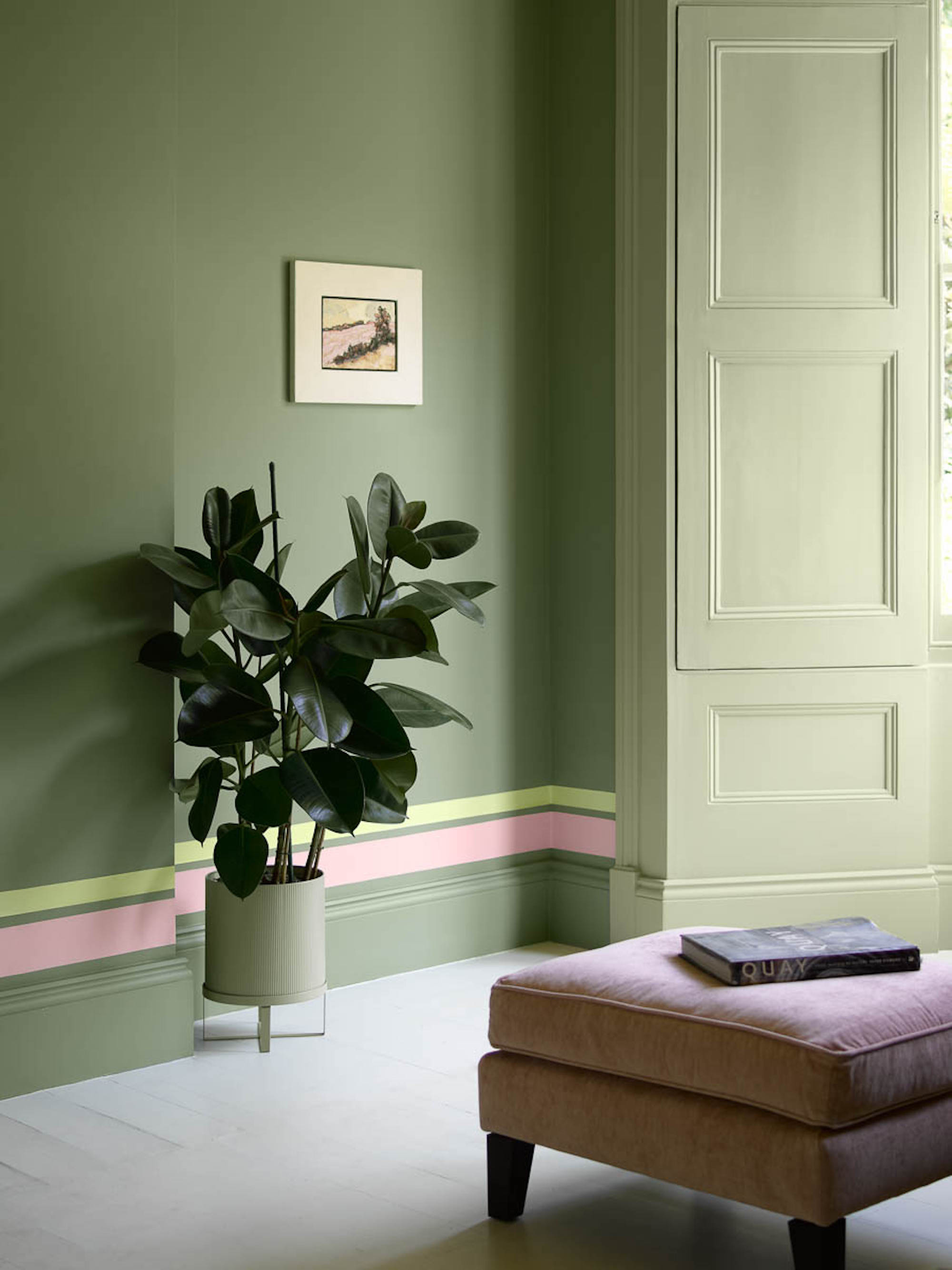 A sage green, colour drenched space with a colourful, striped detailing above the skirting board, and a blush pink foot stool in front.