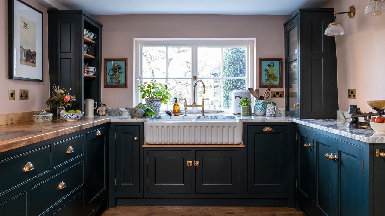 belfast sink in fromt of a window in a small kitchen with poink walls and dark green cabinets