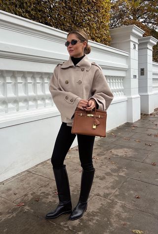 A woman wearing a tan cropped peacoat over a black turtleneck with black leggings, black riding boots, and a brown handbag.