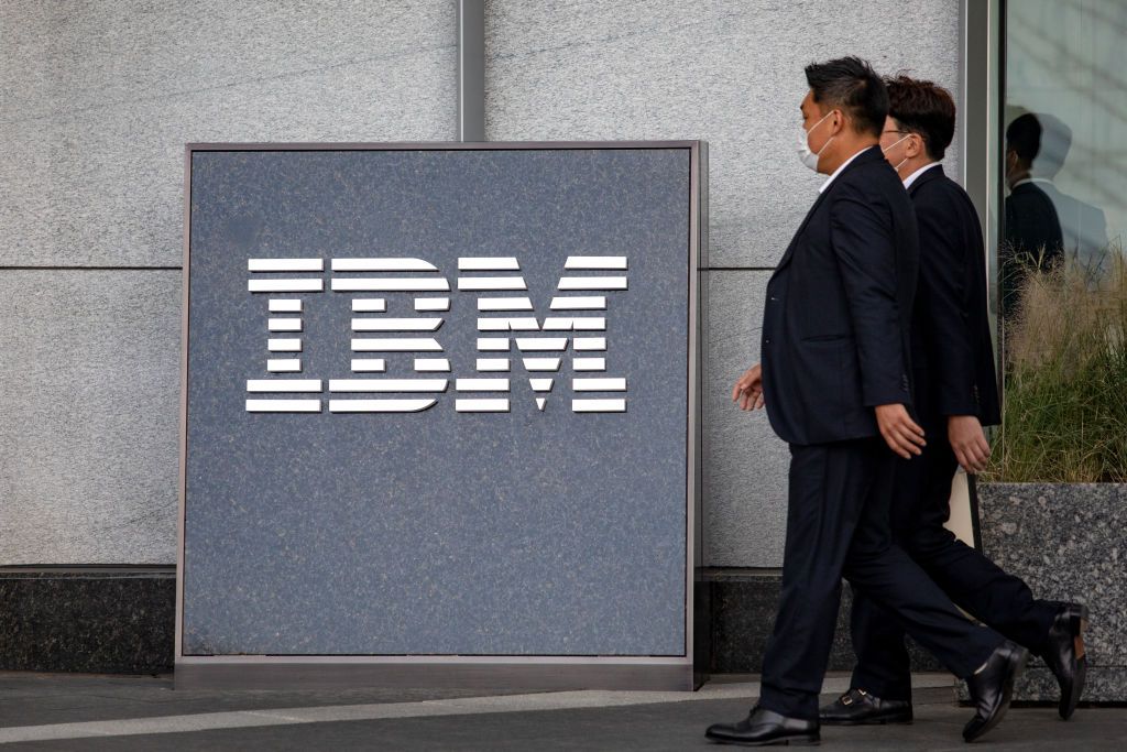 The IBM logo (stylised letters that read &quot;IBM&quot;) on a board against a concrete wall, with two businessmen entering from the right of the frame wearing business suits and medical masks