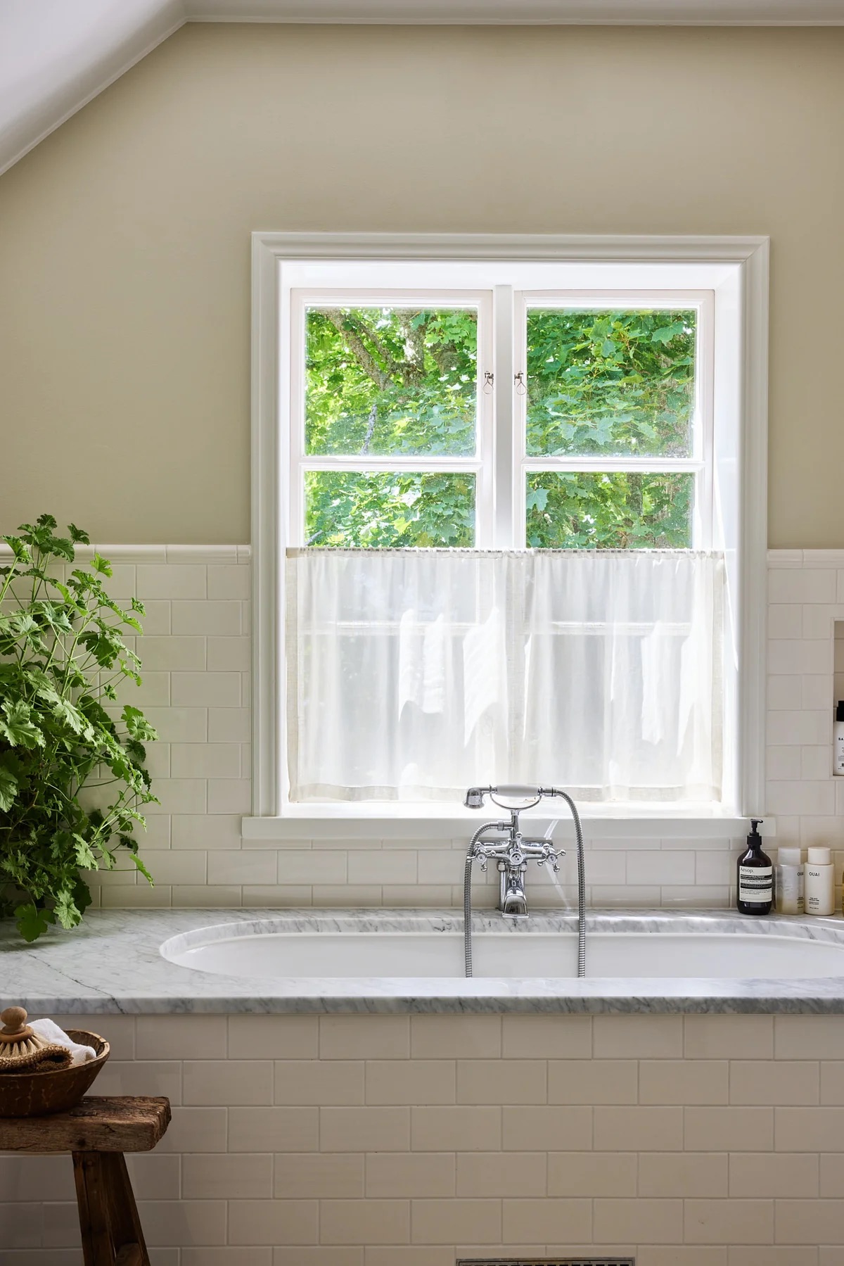 A bathroom with caf&amp;eacute; curtains over built-in bath with subway tiles and marble top, cream walls, and potted plant