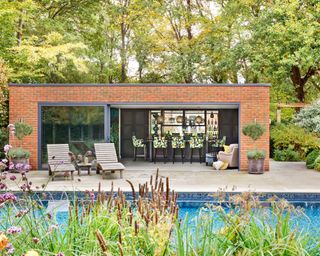 Brick pool house ideas with deck chairs and kitchen area, behind a pool with lush planting around it.