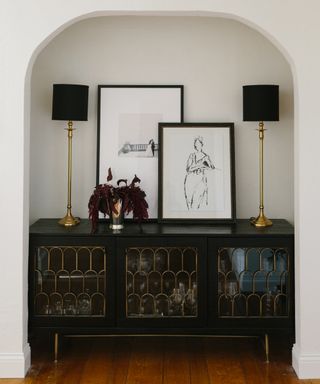 A hallway with recessed alcove filled iwth a black and gold sideboard, gold lamps with black shades, and framed artwork leaning against the wall