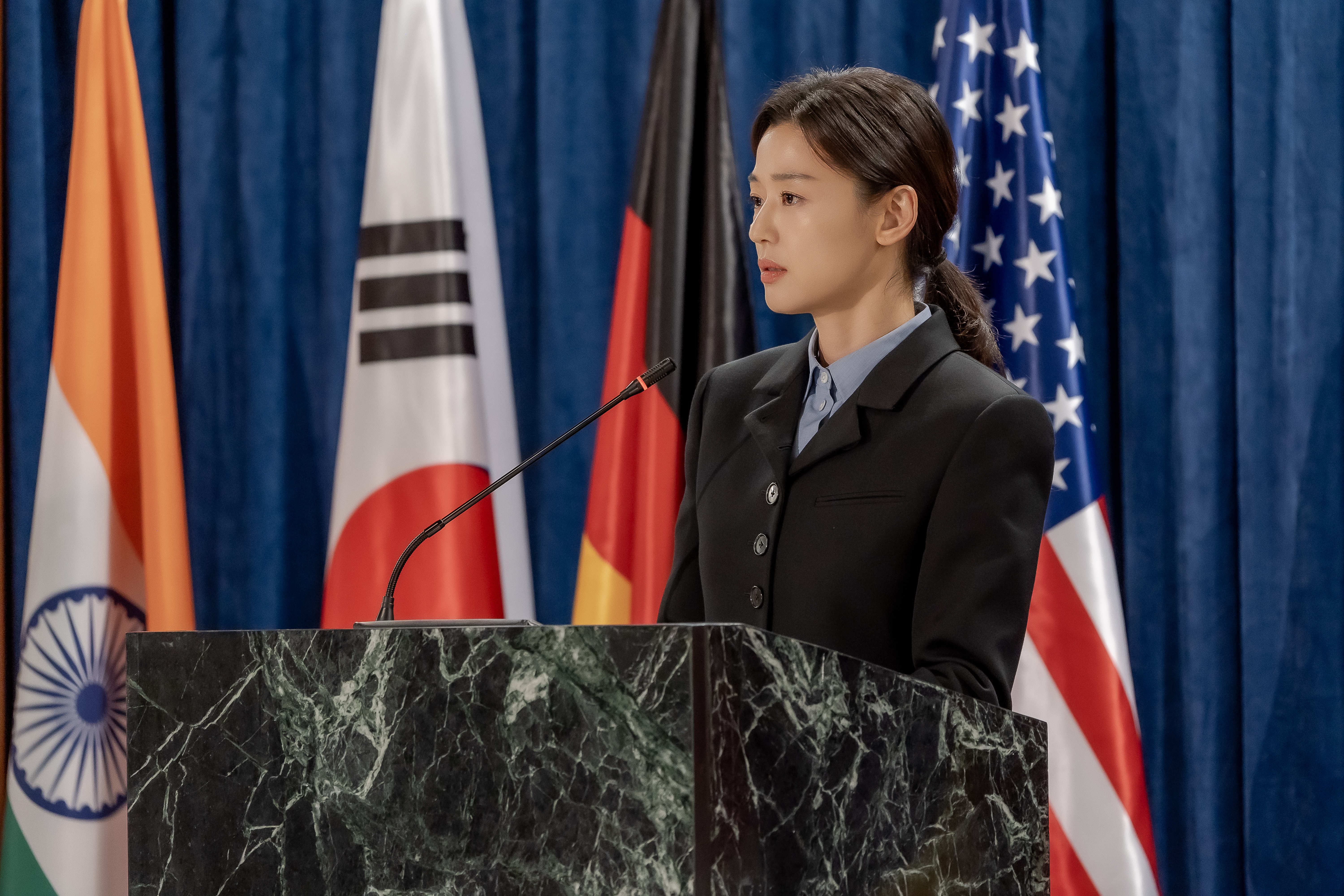 Gianna Jun at a podium in front of flags in &#039;Tempest.&#039;